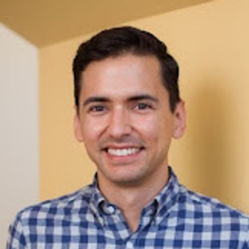 Smiling man with short dark hair wearing a blue-and-white checkered shirt and gray pants, standing with hands in his pockets against a light yellow wall.